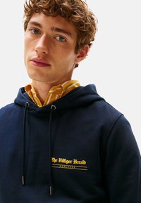 Giovane con capelli ricci che indossa una felpa blu navy sopra una camicia a righe gialla con colletto, il testo sulla felpa recita "The Hilfiger Herald MCMLXXXV".