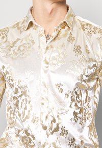 Camisa crema brillante con botones y patrón floral dorado. Textura suave, detalle de cuello y brillo visible de la tela que realzan los elementos del diseño.