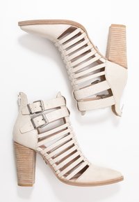 Bottines beige à talons hauts, présentant une texture crocodile, un design ouvert avec plusieurs sangles, des boucles argentées et un talon en bois rayé.