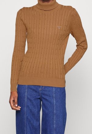 Pull col roulé torsadé marron porté avec un pantalon en jean bleu, la main gauche de la personne posée sur la hanche, la main droite détendue le long du corps.