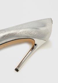 Sølvfarvet stilet sko med tekstureret overflade, der har en spids tå og metallisk detalje på hælen. Glat indre med beige foring.