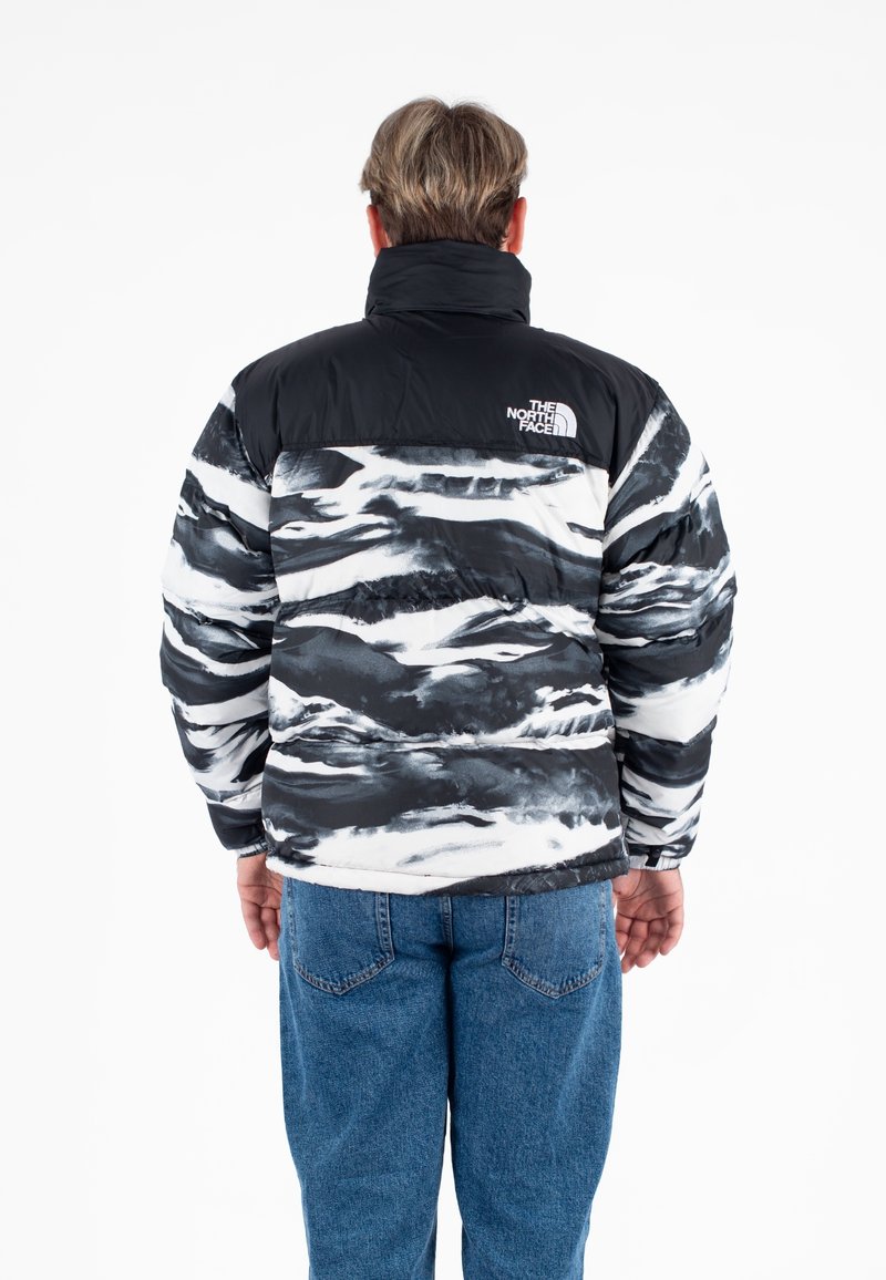 Giacca a vento nera e bianca con motivo astratto a onde, colletto alto e logo sul retro. Indossata con jeans blu. Tessuto morbido e texture.