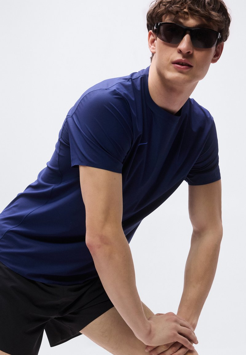 Giovane uomo con occhiali da sole neri, maglietta sportiva blu navy e pantaloncini neri, chino in avanti con le mani sulle ginocchia su uno sfondo semplice.