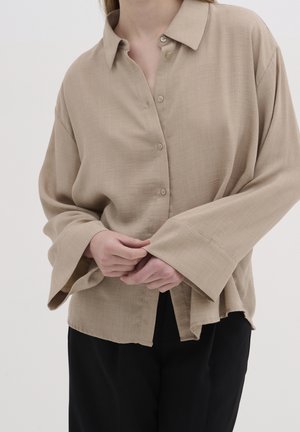 Personne portant une chemise beige ample à boutons avec de larges manches et un pantalon noir, mains doucement jointes à la taille.