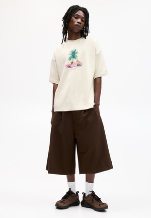Beige oversized t-shirt med en färgglad grafik av en palm och en bil, tillsammans med lösa bruna shorts och bruna sneakers med svarta accenter.