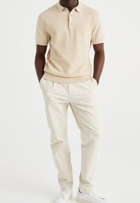 Beige kortärmad pikétröja med ett subtilt texturmönster, i kombination med vita byxor och vita sneakers. Pikétröjan har en tvåknappars hals.