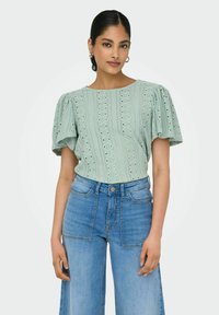 Hellgrüne Bluse mit puffärmel, die ein strukturiertes Muster und Aussparungen aufweist. Kombiniert mit hochgeschnittenen blauen Jeans.