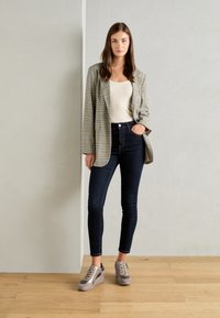 Blazer a cuadros en beige y negro sobre una blusa crema, combinado con jeans skinny oscuros y zapatillas gris claro con cordones. Fondo de madera.