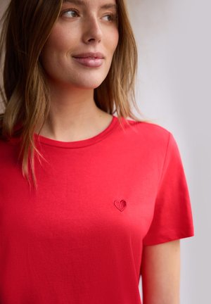 Młoda kobieta w czerwonej koszulce z małym haftowanym sercem na piersi, patrząca lekko w lewo.