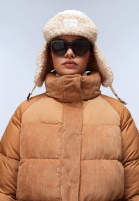 Kurtka puchowa w kolorze piaskowym z żeberkową teksturą z korduroyu i wysokim kołnierzem; zestawiona z kremowym, futrzanym kapeluszem i dużymi, czarnymi okularami przeciwsłonecznymi.