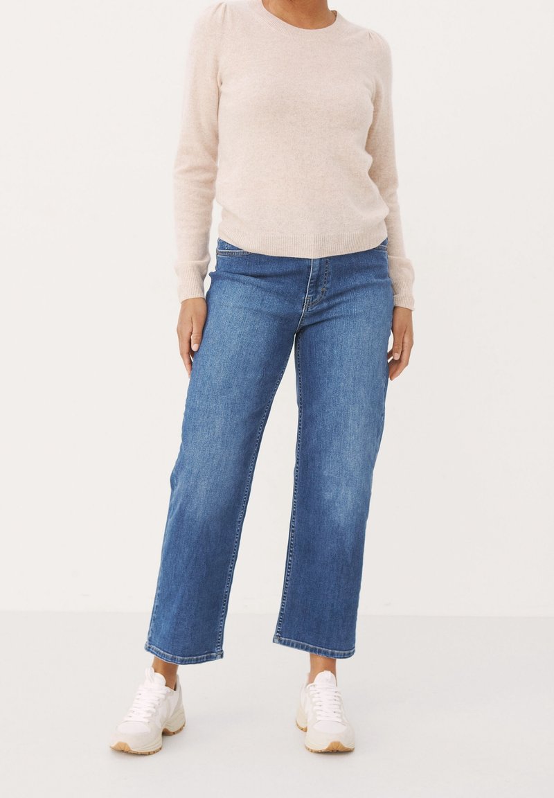 Jasnoróżowy sweter z długim rękawem w połączeniu z niebieskimi dżinsami o średnim stanie i szerokich nogawkach oraz białymi sneakersami. Miękka tekstura; prosty wzór; brak wzorów.