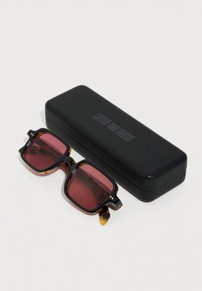 Τετράγωνα γυαλιά ηλίου με καφέ σκελετό τύπου χελώνας και φακούς με κόκκινη απόχρωση, δίπλα σε μία μαύρη υφασμάτινη ορθογώνια θήκη με το λογότυπο «Human Made» χαραγμένο στην κορυφή.