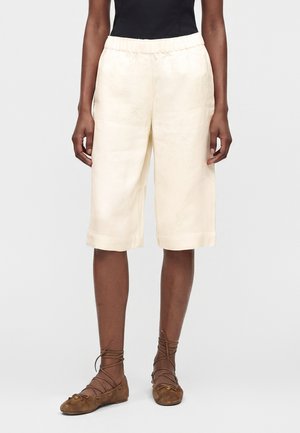 Personne portant un short ample crème jusqu'aux genoux, un haut noir et des chaussures plates à lacets marron, debout devant un fond uni.