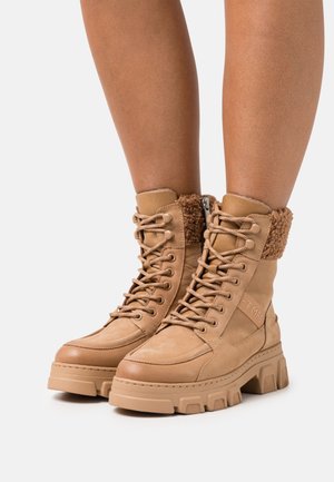 Beige veterlaarzen met stevige zolen en met fleece gevoerde kraag, gedragen op blote benen tegen een effen lichte achtergrond.