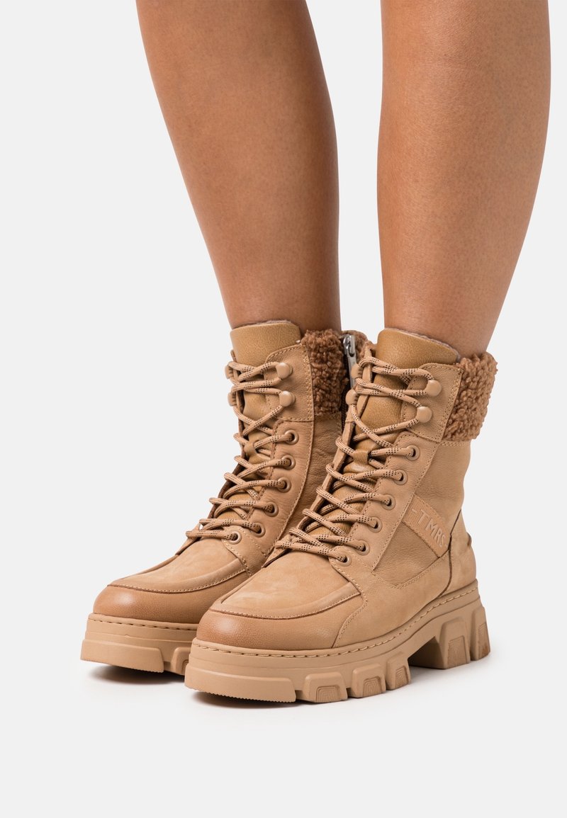 Bottes à lacets couleur tan avec semelles épaisses et cols doublés en polaire, portées sur des jambes nues sur un fond clair uni.