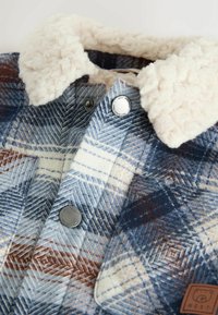 Veste à carreaux dans des tons de bleu, blanc et marron. Élégante avec un col beige et duveteux, des boutons en métal, et un tissu texturé avec un motif en chevrons.