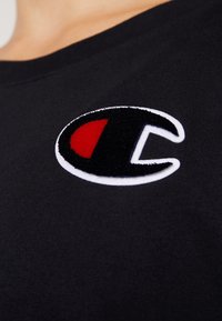 T-shirt nera con un logo circolare ricamato in rosso e bianco. Il tessuto appare liscio con scollatura a girocollo.