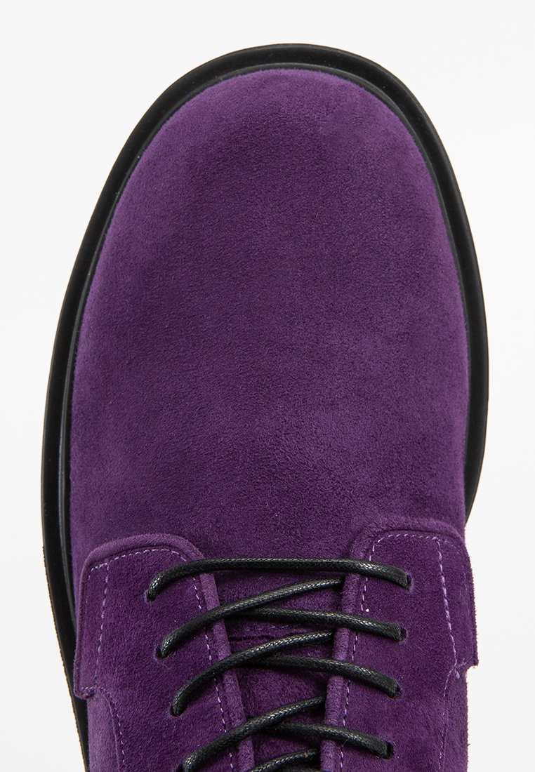 Bottine en daim violette avec semelle noire. Dotée de six œillets noirs et de lacets assortis. Texture lisse sans motifs ni accents visibles.