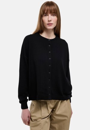 Femme portant un cardigan noir boutonné et un pantalon plissé beige, debout devant un fond clair uni.