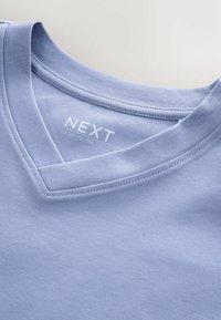 T-shirt en coton bleu clair à col en V, montrant une encolure cousue et une étiquette imprimée "NEXT GROWN ON TEE" à l’intérieur du col.