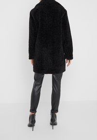 Svart, fluffig, oversized kappa med texturerad yta, långa ärmar och krage. Kombinerad med åtsittande, glansiga svarta byxor och högklackade skor.