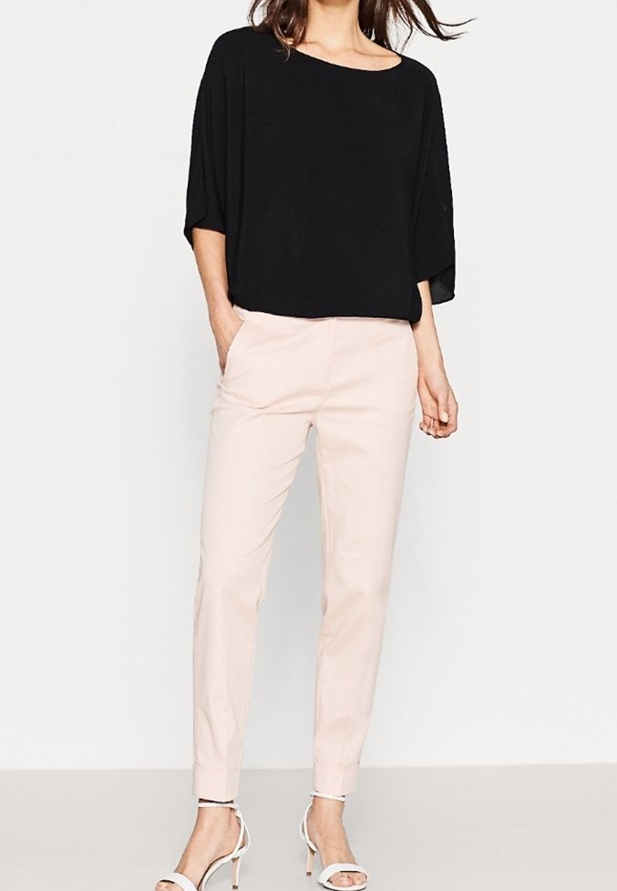 Haut noir ample à manches courtes associé à un pantalon tapered rose clair et des talons blancs à lanières, sur fond uni.