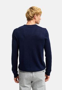 Maglione lavorato a maglia blu navy con maniche lunghe e collo tondo, caratterizzato da una texture liscia e un design aderente. Indossato sopra pantaloni grigi.