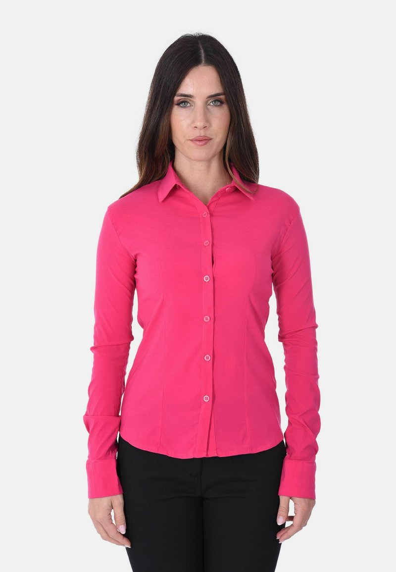 Donna con lunghi capelli scuri che indossa una camicia rosa vivace con bottoni e pantaloni neri, in piedi su uno sfondo bianco semplice.