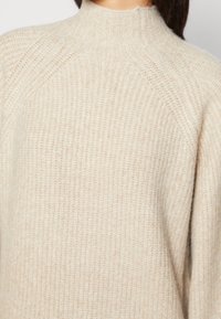 Pull tricoté couleur crème avec un col montant, un motif texturé et des manches raglan, fabriqué dans un matériau doux et chaud.