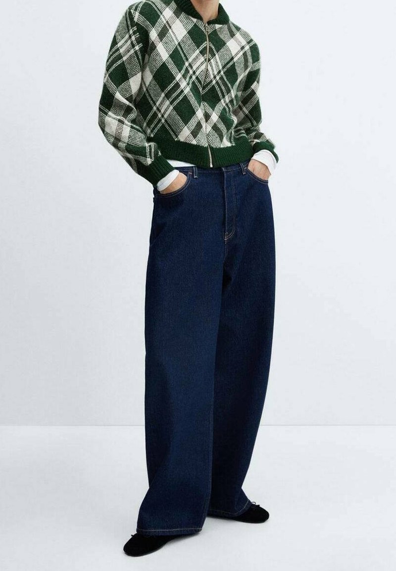 Pull zippé à carreaux verts et blancs avec un ourlet côtelé, associé à un jean denim bleu foncé à jambes larges. Le mannequin pose les mains dans les poches.