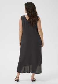 Femme avec de longs cheveux ondulés portant une robe noire sans manches, ample et longueur cheville, ainsi que des sandales à talons noires, de dos.