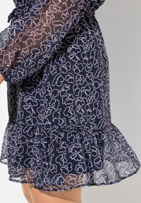 Dunkelblaues Kleid mit semi-sheer Textur, verziert mit rosa Blumenmustern und einem volanten Saum. Lange Ärmel mit elastischen Bündchen.