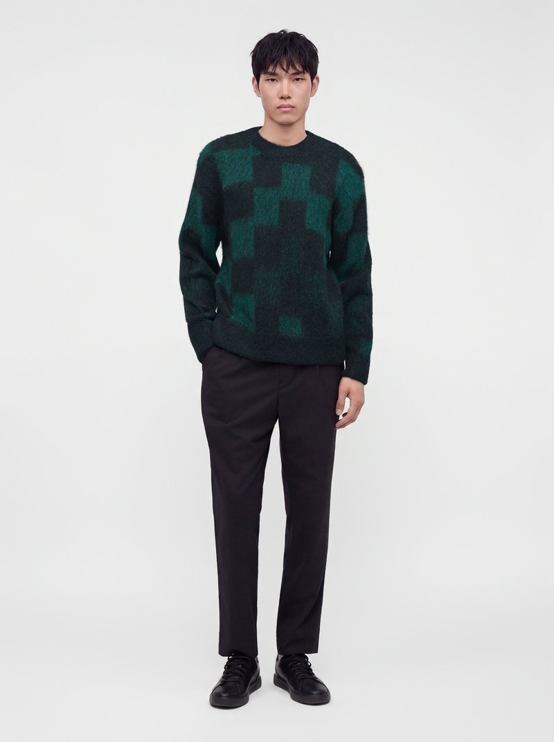 Homme debout, les mains dans les poches, portant un pull à motifs vert foncé et noir, un pantalon noir et des chaussures noires sur fond blanc.