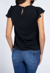 Blusa negra de manga corta con mangas con volantes y diseño de abertura en la espalda, combinada con jeans azules que tienen un acento de logo triangular en el bolsillo.