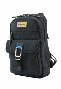 Zaino in tessuto nero con tasca anteriore a pattina, dettagli in hardware blu e oro, e una patch con logo giallo e rosso. Presenta spallacci imbottiti.
