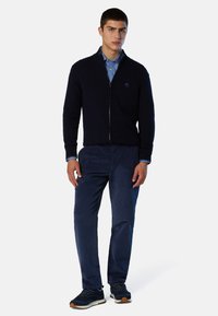 Maglione blu navy con zip e polsini a coste, abbinato a pantaloni di velluto a coste blu navy e sneakers blu navy. Camicia a quadretti blu sotto.