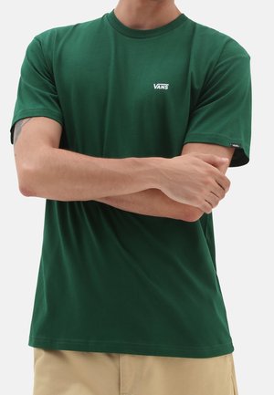 Person trägt ein grünes Vans T-Shirt mit kleinem Logo auf Brust und Ärmel, die Arme verschränkt, beige Hosen unterhalb der Taille sichtbar.