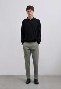 Polo negro de manga larga con cuello, combinado con pantalones chinos verde claro y zapatos negros. Diseño sencillo, estilo minimalista.