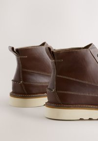 Ruskeat nahkaiset nilkkurit, joissa on valkoinen kontrastitikkaus ja beige pohja. Kengissä on kaareva muotoilu ja vetolenkit takana.