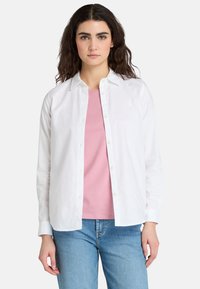 Camisa blanca de botón hecha de algodón, con mangas largas y corte recto, llevada sobre una camiseta rosa. Unos pantalones vaqueros de mezclilla azul completan el atuendo.