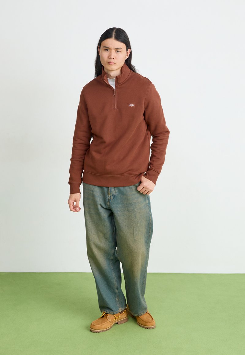 Felpa zip beige con un piccolo logo, abbinata a jeans oversize di un azzurro chiaro e zoccoli color cammello su un pavimento verde.