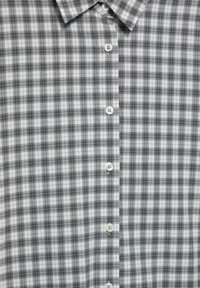 Chemise à carreaux gris et blanc avec boutons, col pointu et six boutons blancs visibles sur le devant.