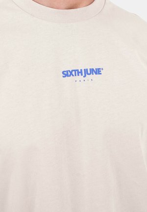 Kremowy T-shirt z niebieskim tekstem "SIXTH JUNE" i mniejszym słowem "PARIS" nadrukowanym na piersi pod dekoltem.