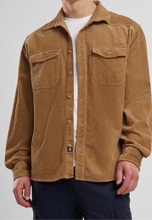 Hombre con una camisa marrón de pana abotonada sobre una camiseta blanca, combinada con pantalones oscuros, de pie frente a un fondo gris liso.