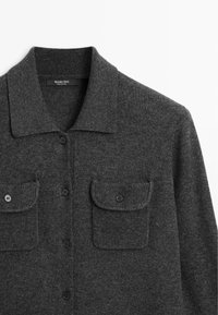 Chemise tricotée gris foncé avec col, boutonnée sur le devant, et deux poches poitrine à rabat avec bouton, posée à plat sur fond blanc.