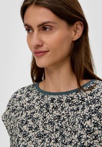 Blumenmusteroberteil in Creme und Dunkelblau, mit einem Rundhalsausschnitt und kontrastierendem Türkisbesatz sowie kurzen Ärmeln. Weiche Stofftextur.