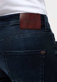 Jeans din denim albastru închis au un patch din piele maro pe talie și un accent metalic pe buzunarul din spate, cu cusături decorative.
