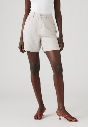 Mørkhudet person iført hvide cowboyshorts med høj talje, hvid ærmeløs top og røde sandaler med lav hæl står på grå baggrund.