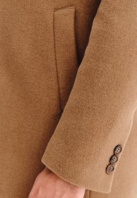 Rękaw brązowego wełnianego płaszcza o gładkiej fakturze, wykończony trzema ciemnymi guzikami i dyskretnym detalem kieszeni.