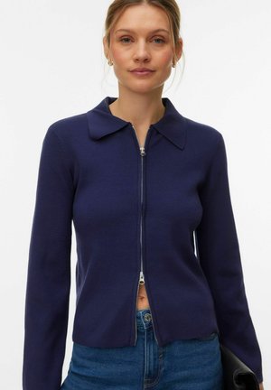 Cardigan marine à fermeture éclair avec col, en matière tricotée côtelée. Comprend des manches longues et une silhouette ajustée. Mis en valeur par une fermeture éclair argentée.